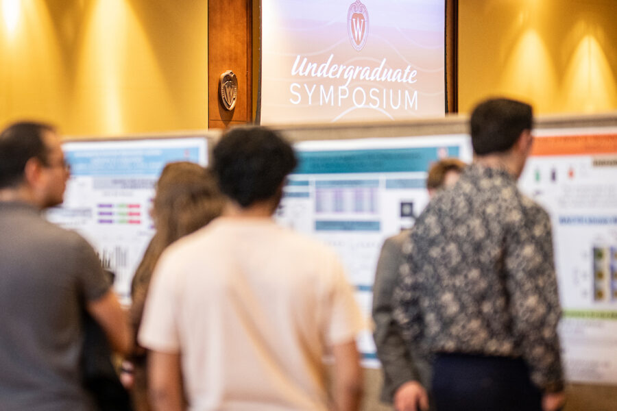 the focus of this image is on the background, with research posters tacked to boards and a sign above that says "undergraduate symposium." The blurry foreground shows attendees of the symposium looking at a poster. their backs are all to the camera