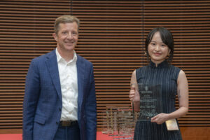 two people stand smiling, the woman on the right holds a glass plaque