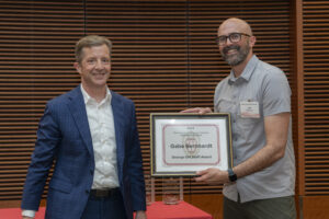 two men smile, one holds a framed certificate