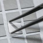 A pair of tweezers lays a square piece of glass in an array of other square pieces of glass